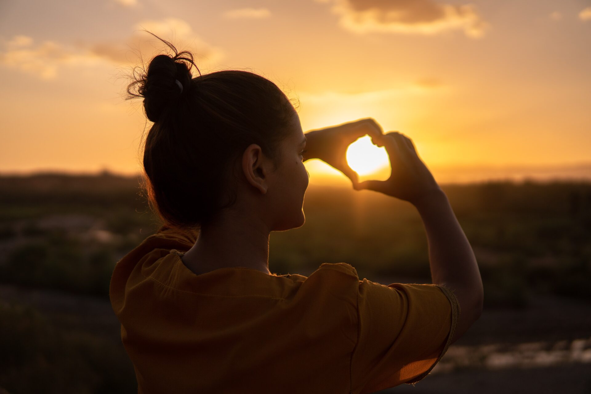 A lady holding up a heart in front of a sunset.